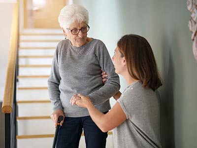 services 2 A caring woman assisting an elderly lady up the stairs, providing support and ensuring her safety.